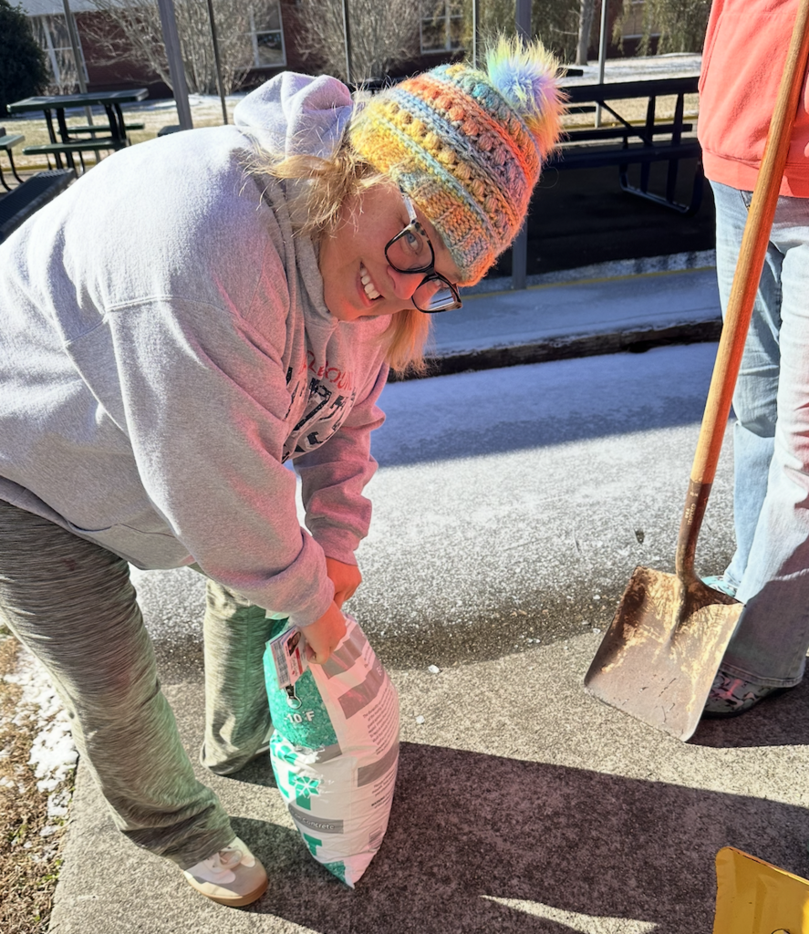 School staff member wearing a winter hat and glasses pours ice melt from a bag onto an icy sidewalk near outdoor seating at a school.