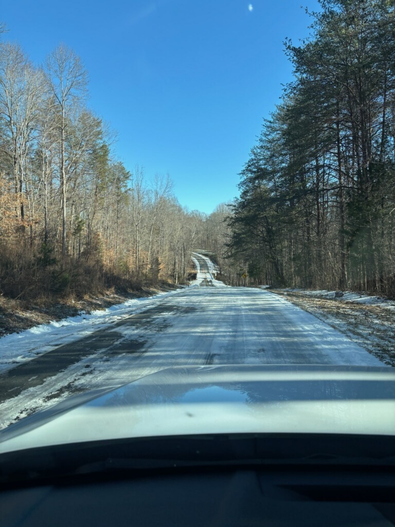 Narrow rural road covered in ice and packed snow, stretching into the distance between wooded areas, photographed from inside a vehicle with the hood visible at the bottom of the frame.