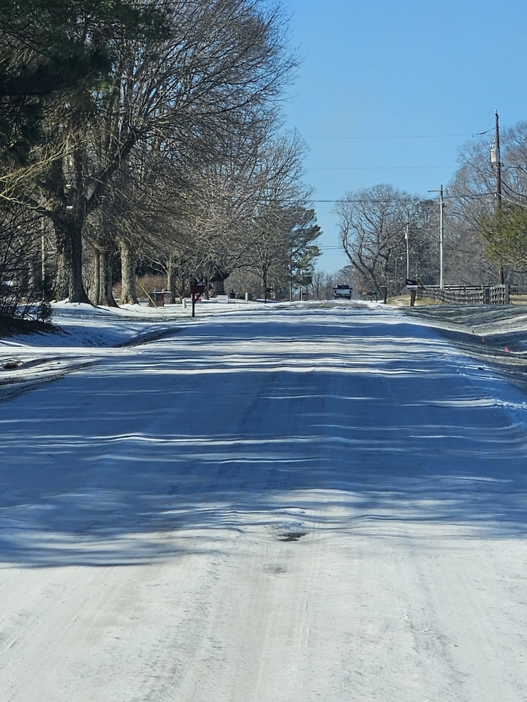 Residential road coated with ice and snow, lined with trees, mailboxes, and utility poles, with long icy patches across the roadway and a clear blue sky overhead.