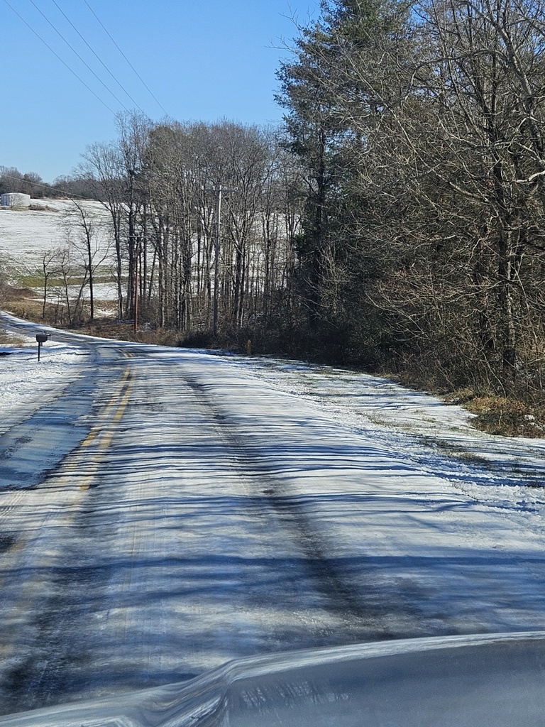 Icy two-lane rural road viewed from a vehicle, with snow and ice covering the pavement, bare trees lining both sides, and open fields in the distance under a clear blue sky.