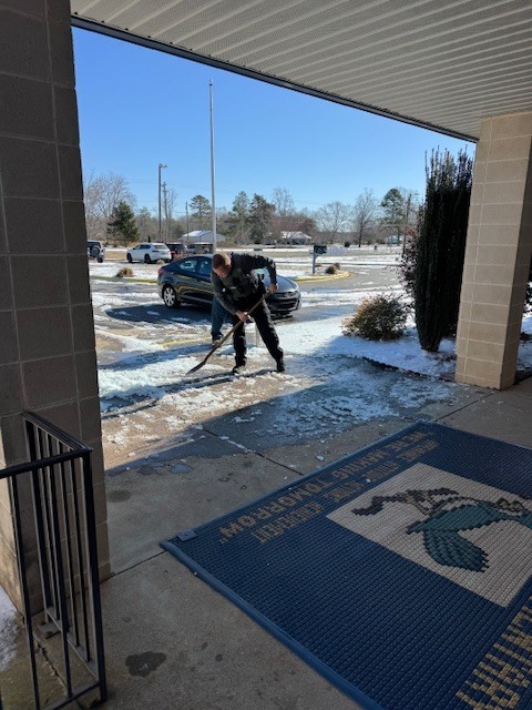 An adult uses a shovel to break up ice on a school sidewalk near the front entrance, with parked cars and patches of snow visible in the background.