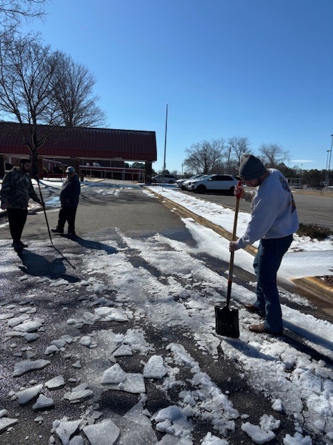 Three adults work together to shovel and move ice from a school parking lot, with large broken pieces of ice scattered across the pavement.