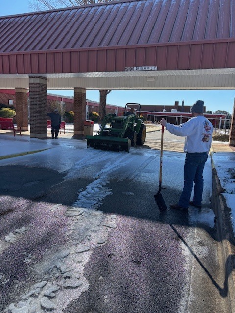 A green tractor clears ice from a covered school drop-off area while an adult with a shovel stands nearby, guiding the cleanup on a sunny winter day.