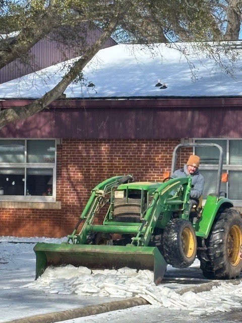 An adult drives a small green tractor with a front blade, pushing chunks of ice off a school walkway in front of a brick building.