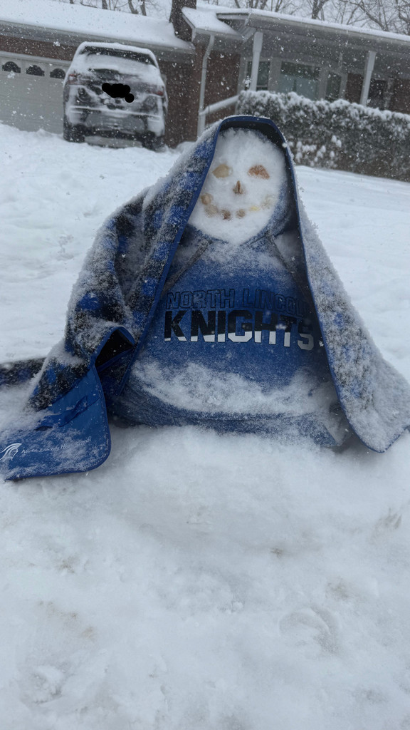 A snowman sits in a snowy driveway wrapped in a blue “North Lincoln Knights” hoodie. Snow covers the ground and a parked vehicle is visible in the background.