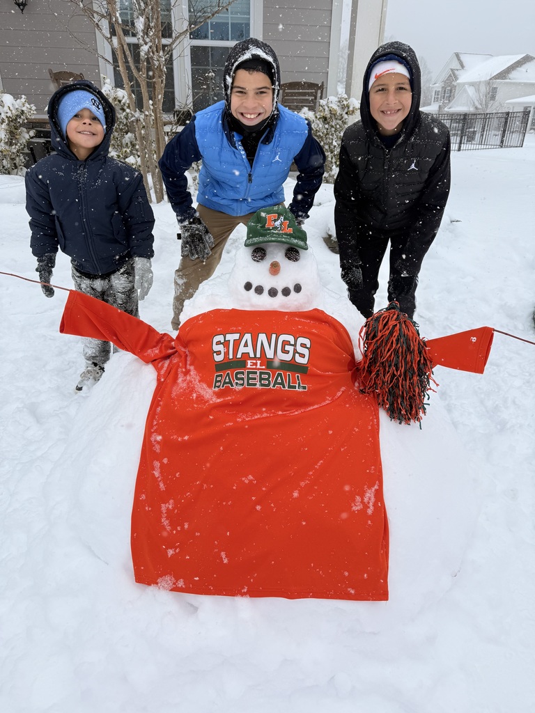 Three children stand in the snow behind a decorated snowman wearing a red “STANGS EL Baseball” shirt and a green hat. The snowman has outstretched arms, and the children smile in a snowy front yard.