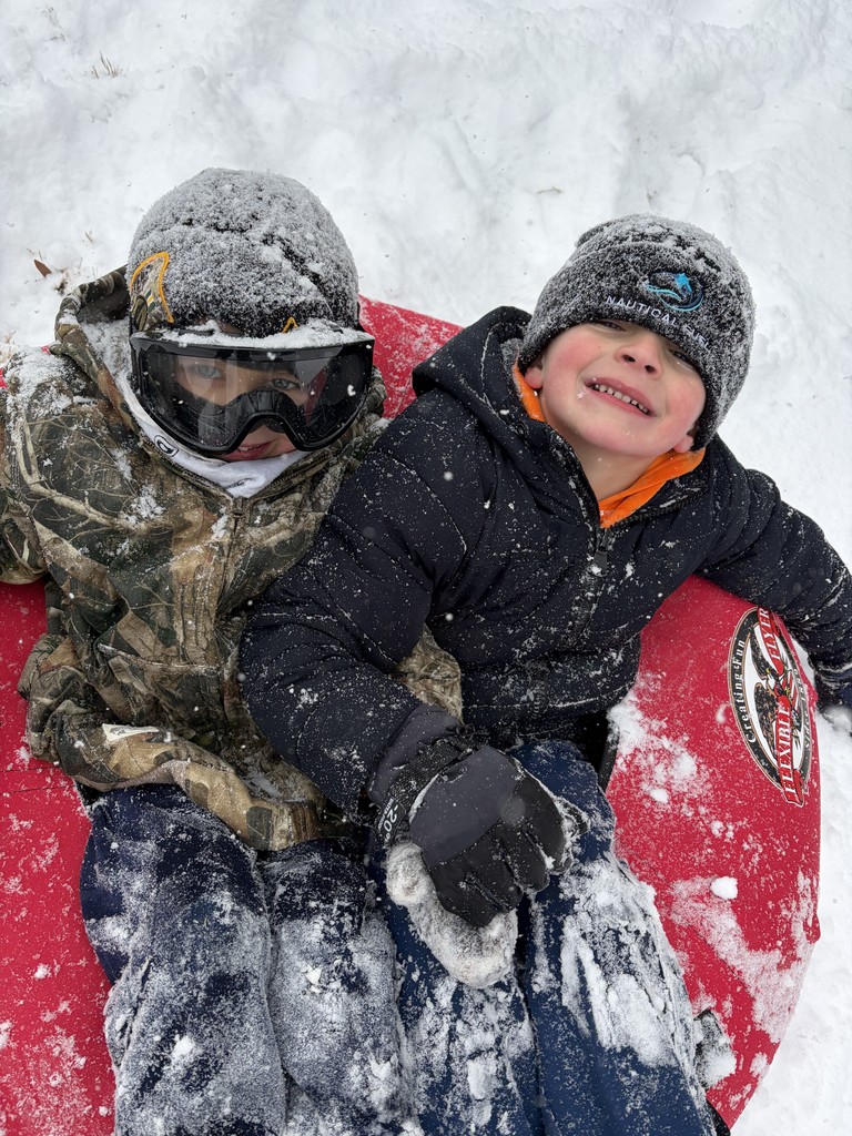 Two children bundled in winter coats and hats sit on a red inflatable sled in fresh snow. One child wears ski goggles and camouflage outerwear, while the other smiles at the camera with snow on their jacket.