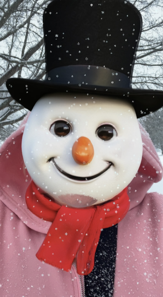 A close-up of a person wearing a snowman face mask with a black top hat and red scarf, standing outdoors as snowflakes fall.