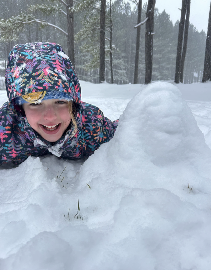 A child in a colorful winter jacket lies on their stomach in the snow, smiling at the camera next to a small mound of snow, with pine trees visible behind them.