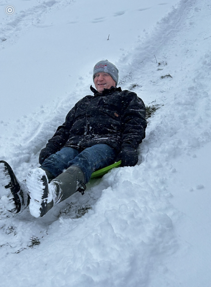 An adult sits on a small sled at the top of a snowy slope, smiling while wearing a winter coat, jeans, boots, and a knit hat, surrounded by packed snow.