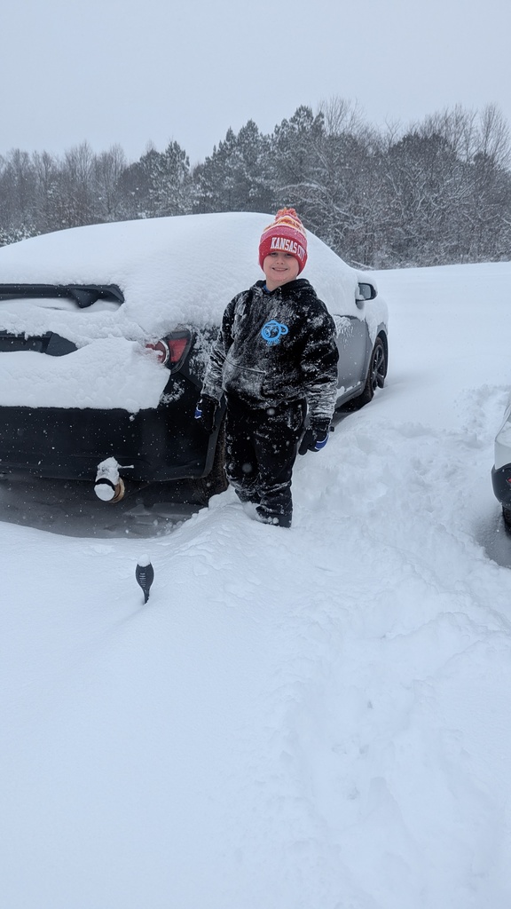 A child wearing a red knit hat and black snowsuit stands smiling in deep snow beside a car covered in fresh snowfall, with trees and falling snow in the background.