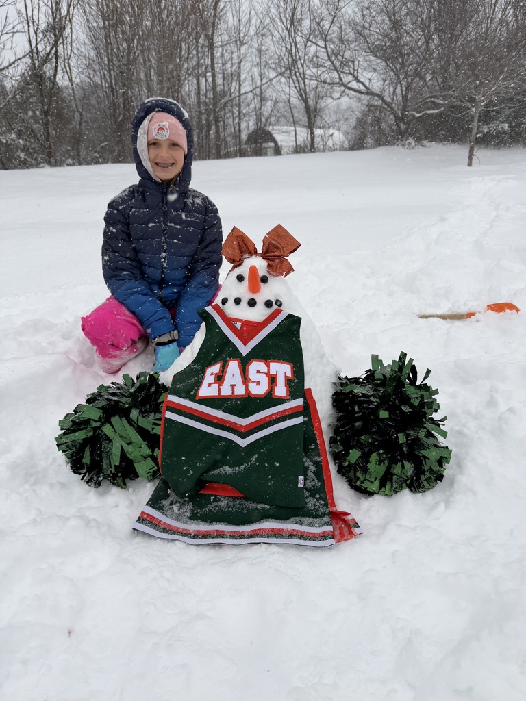 A child kneels in the snow next to a decorated snowman dressed in a green cheer uniform with the word “EAST,” holding green pom-poms, while snow falls in a wooded area.