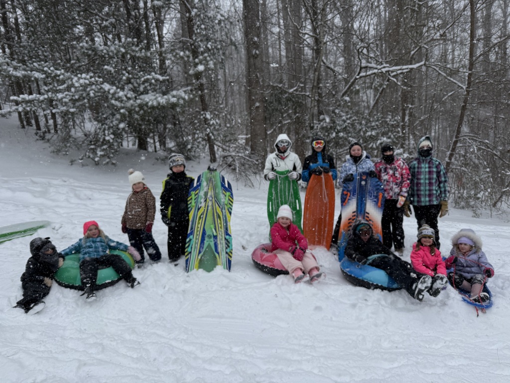 A large group of children bundled in winter coats pose in the snow holding sleds and snow tubes, lined up at the edge of a wooded hill during active snowfall.