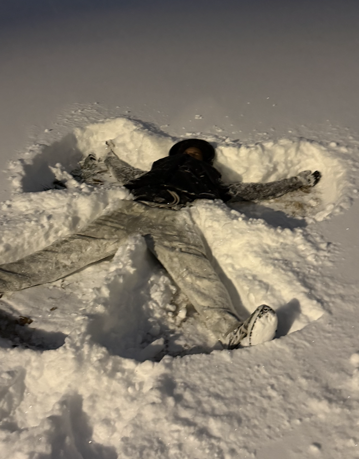 A person lies on their back in deep snow at night, arms and legs spread wide, forming a snow angel. Snow-covered houses and a streetlight are visible in the background.