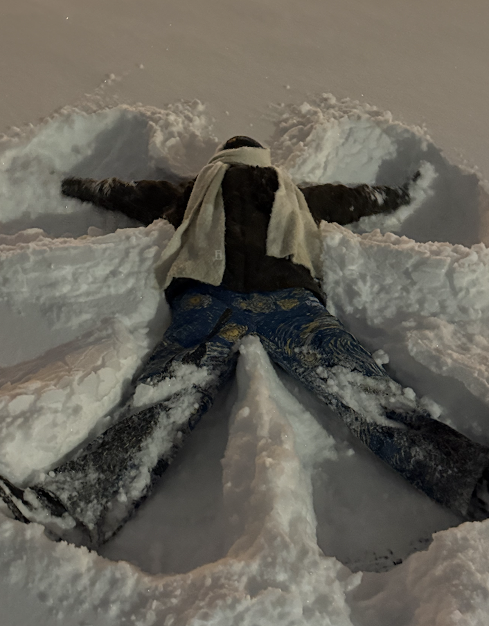 A second nighttime photo of a person making a snow angel, wearing jeans, a dark jacket, and a light-colored scarf. The snow angel shape is clearly visible around their body.