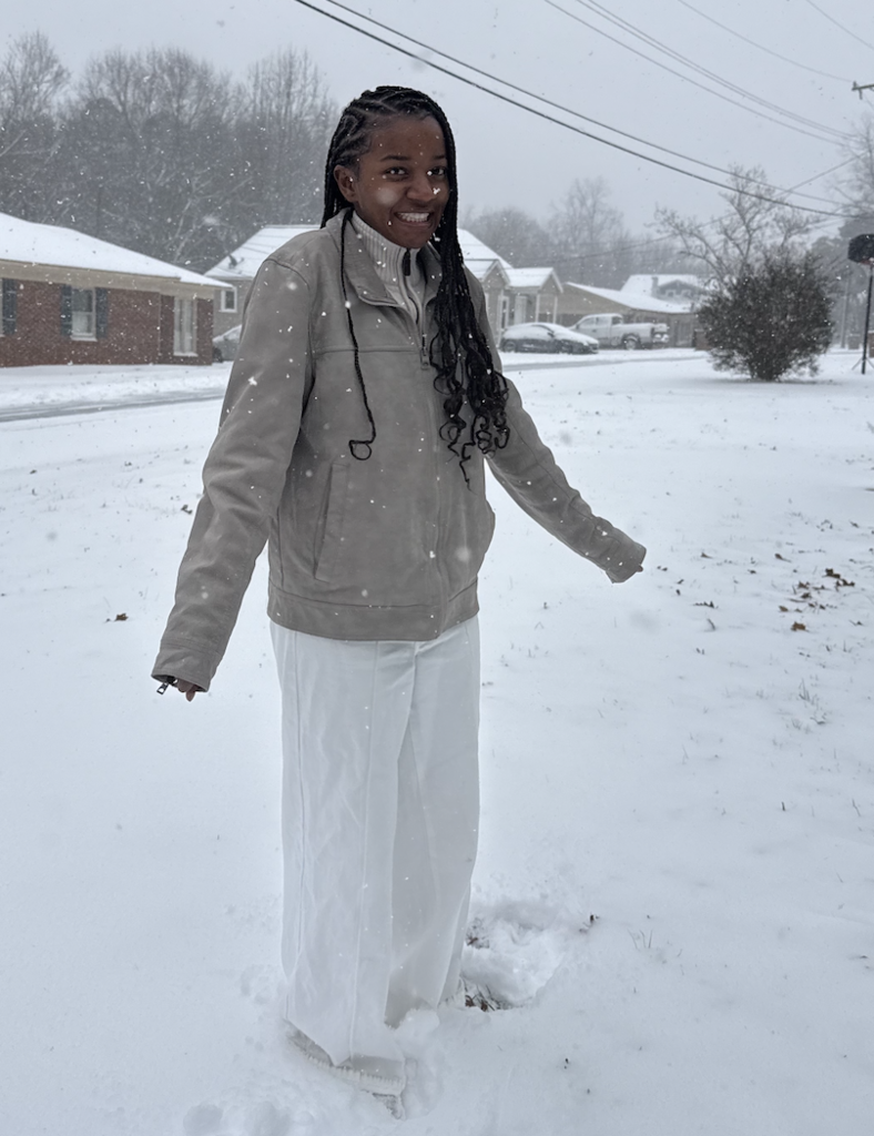 A person stands in fresh snowfall on a residential street during active snow flurries, wearing a tan jacket and white pants. Snow covers the road, rooftops, and lawns around them.