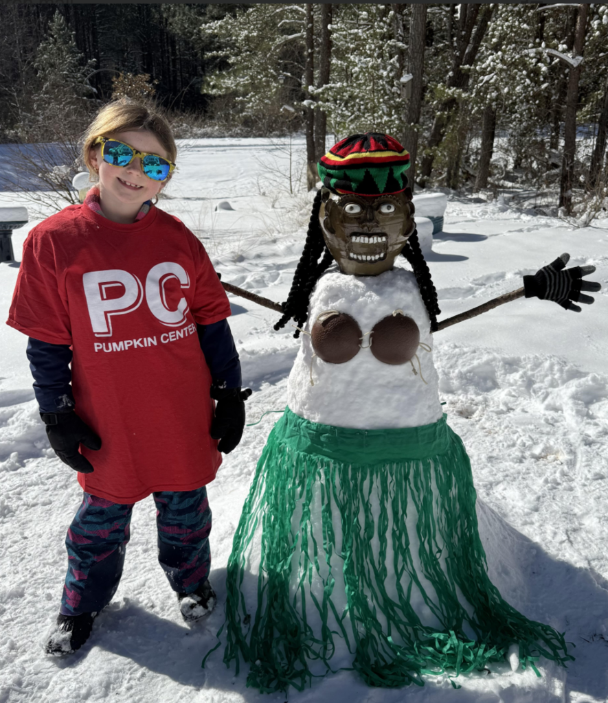 A child wearing a red “Pumpkin Center” shirt and reflective sunglasses stands next to a creatively styled snowman with a decorative mask, braided hair, coconut shell top, and green grass skirt. Both are in a snowy yard.