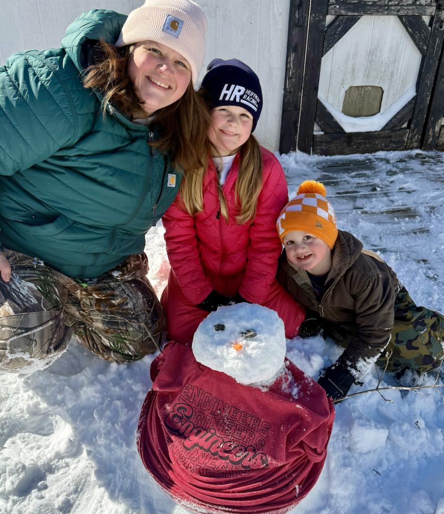 Two children and an adult kneel in the snow around a snowman wearing a red shirt with “Engineers” printed on the back. All three are smiling, dressed in winter clothing, with snow covering the ground and a shed behind them.