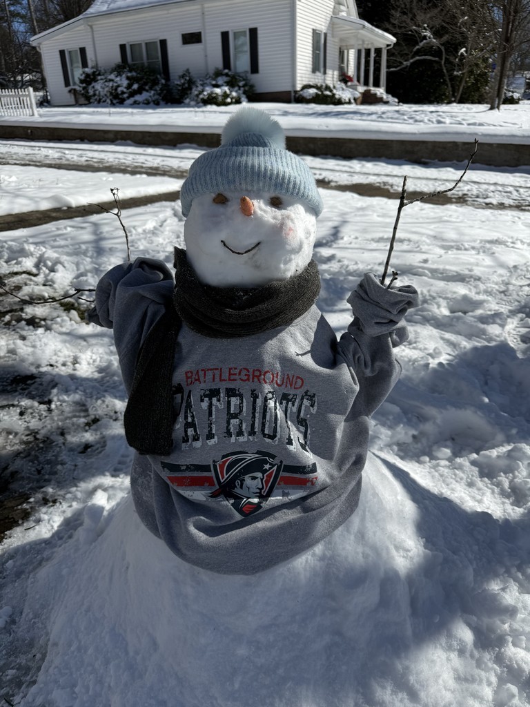 A snowman wearing a light blue knit hat and a gray “Battleground Patriots” hoodie stands in a snowy front yard, holding stick arms up like a celebration. A white house and snow-covered ground are visible behind it.