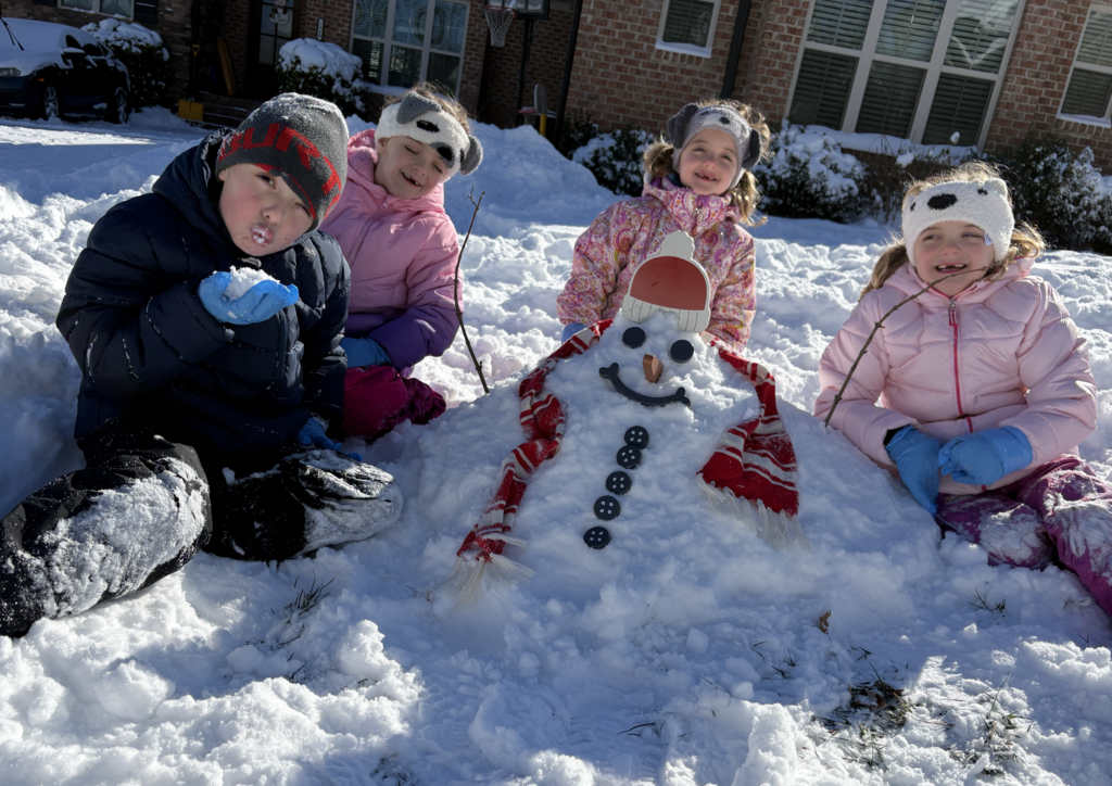 Four young children sit in the snow smiling beside a snowman decorated with a red-and-white scarf, buttons, and a hat. Snow blankets the ground and nearby homes.
