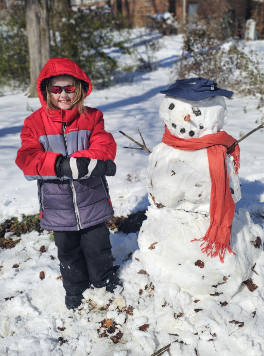 A child in a red winter jacket and sunglasses stands next to a snowman wearing a blue hat and red scarf. Leaves are mixed into the snow around the snowman.