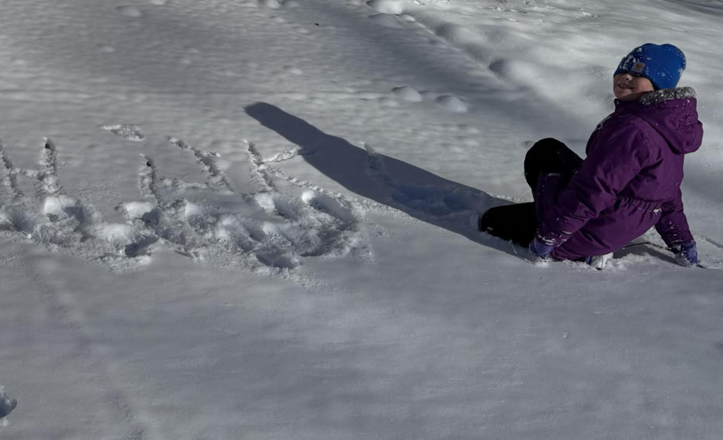 A child sits in the snow beside letters written in the snow. The child wears a purple winter coat and a blue hat, with long shadows cast across the snow.