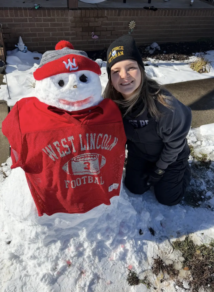 A child kneels beside a snowman wearing a red football jersey reading “West Lincoln Football” and a red knit hat. Snow covers the ground and a brick building is visible behind them.