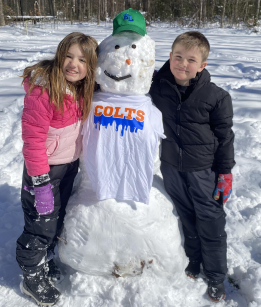 Two children stand beside a snowman wearing a white shirt that reads “COLTS” and a green hat. All three face the camera in a snow-covered yard.