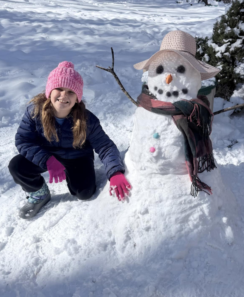 A child wearing a pink knit hat and winter gloves kneels beside a snowman decorated with a wide-brimmed hat, scarf, stick arms, and colorful buttons. Fresh snow covers the yard.