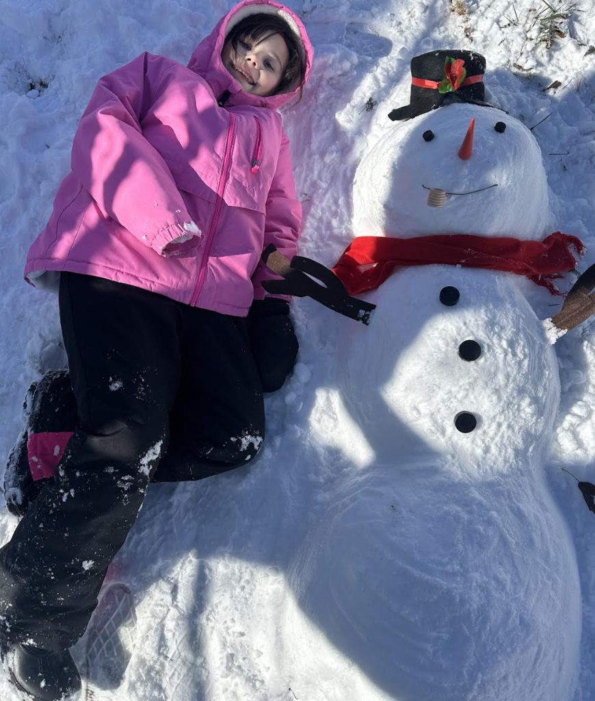 A child lies in the snow beside a snowman wearing a black top hat and red scarf. The child is dressed in a pink winter coat and black snow pants.