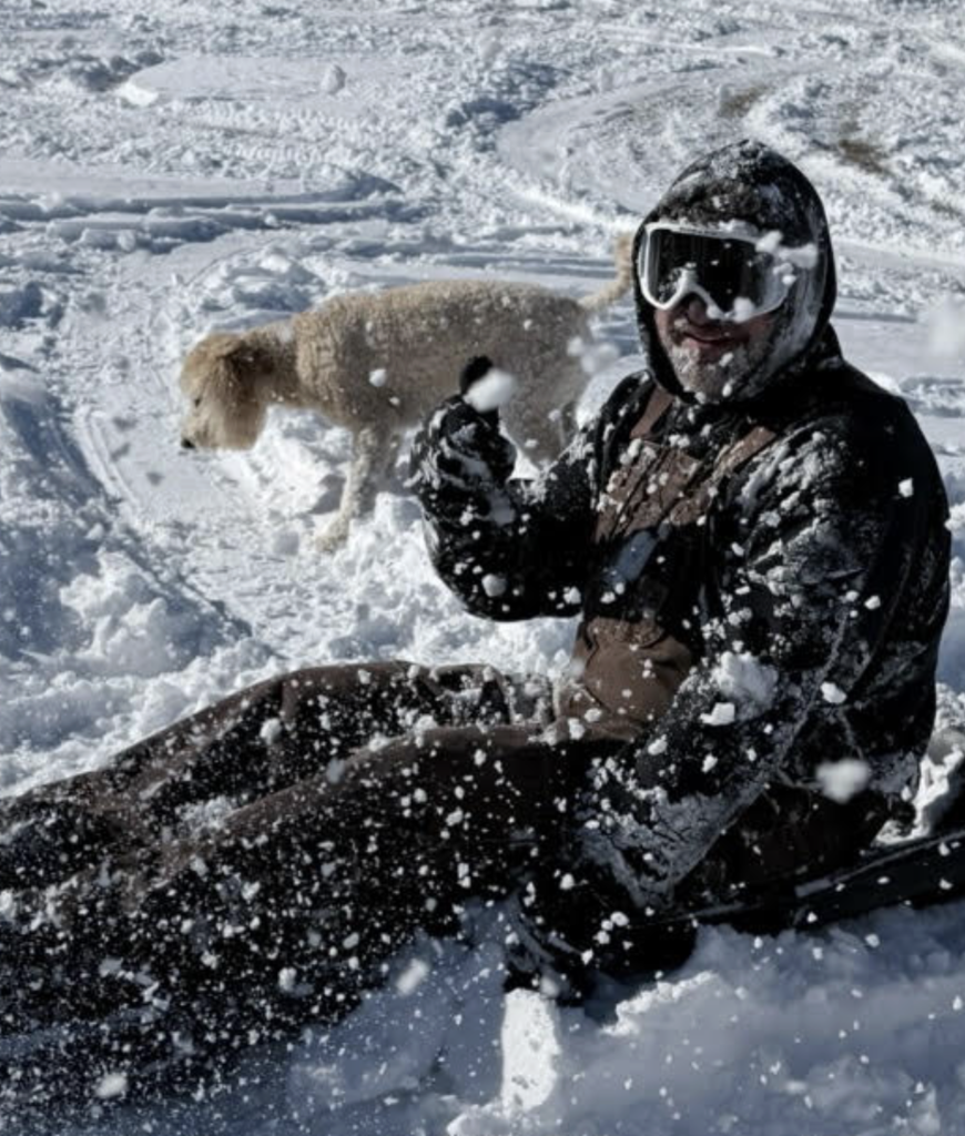 A person wearing winter clothing and snow goggles sits in the snow while tossing snow into the air. A light-colored dog stands nearby in the snowy background.