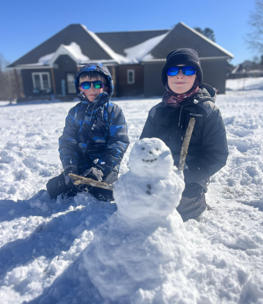 Two children sit in the snow behind a small snowman with stick arms. Both children wear winter jackets, hats, and sunglasses, with a house visible in the background.