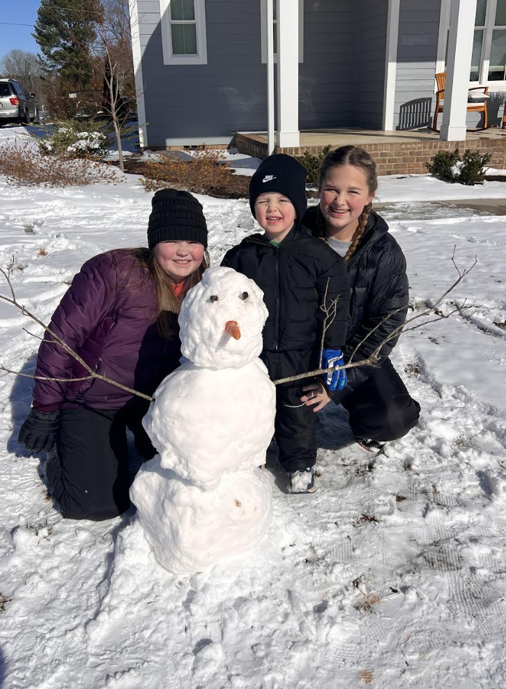 Three children kneel behind a classic three-tier snowman in a sunny yard. The snowman has a carrot nose and stick arms. The children are dressed in dark winter coats, gloves, and knit hats, smiling at the camera.