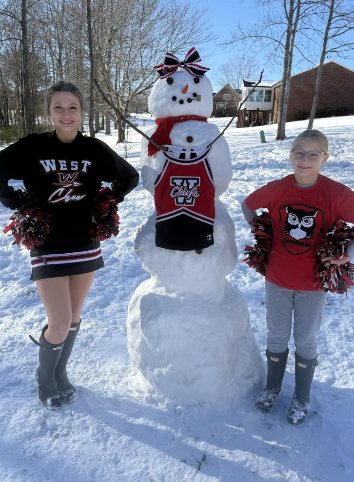 Two students stand beside a tall snowman in a sunny neighborhood yard. The snowman is decorated with a red scarf, a cheer bow on its head, stick arms, and a West Chiefs cheer uniform draped on its front. Both students wear winter boots and hold red and black pom-poms while smiling beside their creation, with snow-covered ground and houses in the background.