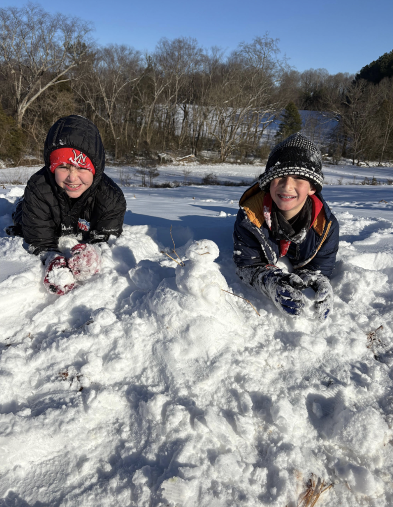 Two children lie on their stomachs in a snowy field beside a small snowman made from three stacked snowballs with stick arms. Both children wear winter coats, hats, and gloves, with snow on their sleeves and hands. A tree line and blue sky appear behind them.