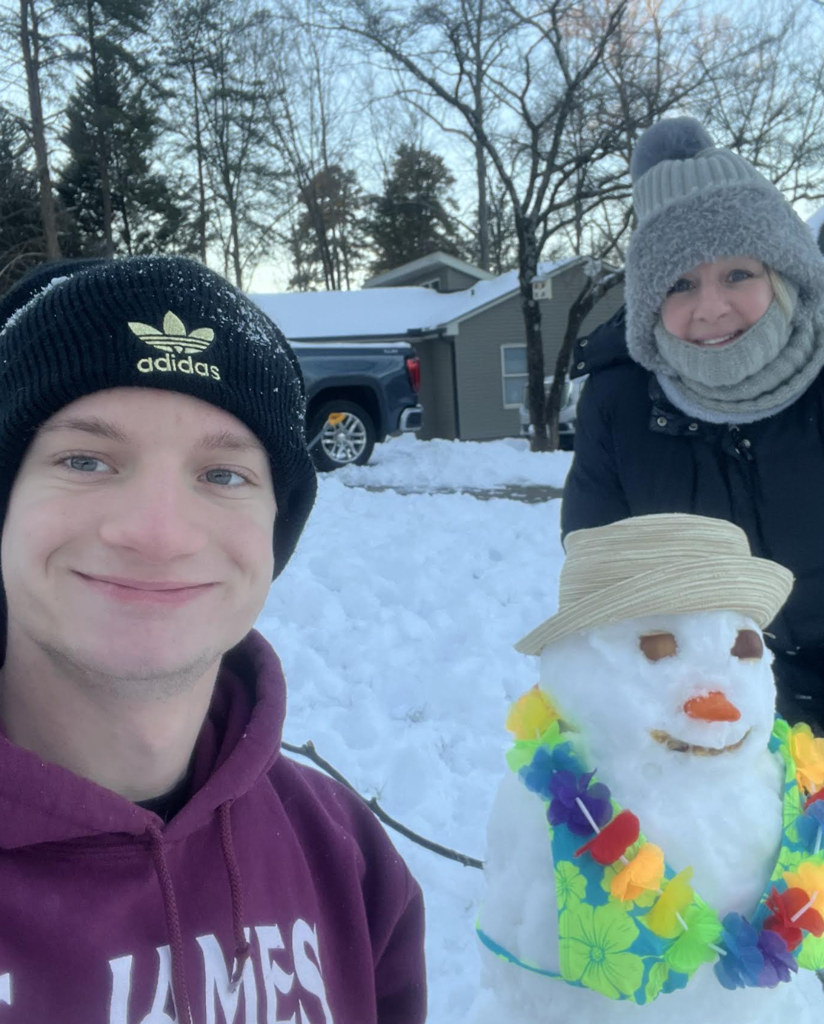 A teenager and an adult stand outdoors with a decorated snowman. The snowman wears a straw hat and a bright tropical lei. Both people are dressed in winter coats, hats, and scarves, with a snowy neighborhood street behind them.