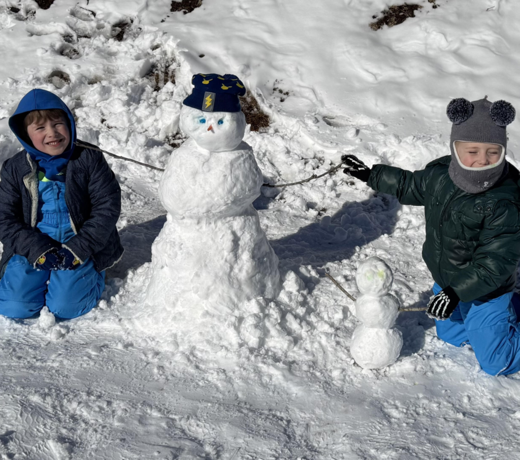 Two young children kneel in the snow beside a medium-sized snowman. The snowman wears a blue winter hat with a lightning bolt design and has blue eyes and a small carrot nose. A smaller snowman stands beside it. The children are bundled in winter coats, gloves, and hats, smiling in bright sunlight.