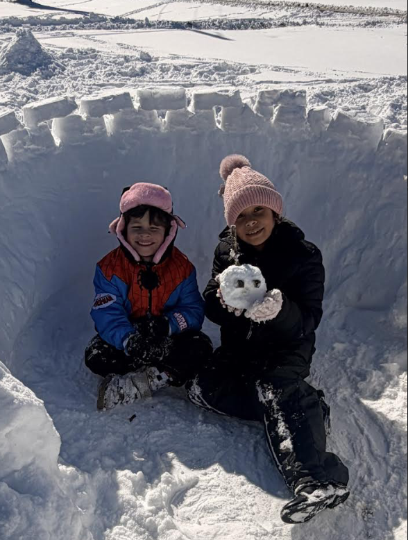 Two children sit inside a snow fort carved into a tall snow wall. One child wears a Spider-Man style winter coat and a pink hat, and the other wears a black coat and pink knit hat while holding a small snowball with pebble eyes.