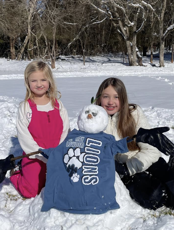 Two children kneel beside a snowman wearing a blue “Lions” T-shirt. The snowman has a carrot nose, pebble eyes, and stick arms. The children wear winter coats and gloves in a sunny snow-covered field.