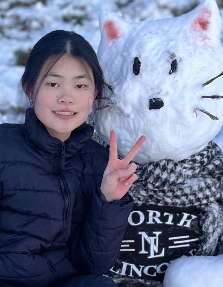 A student poses beside a snow sculpture shaped like a cat wearing a North Lincoln shirt and scarf. The student makes a peace sign while standing next to the snow figure in a winter setting.