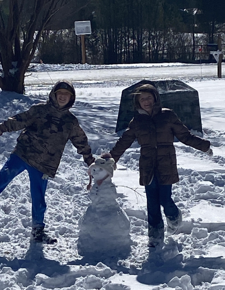 Two children stand beside a tiny snowman in a snowy yard near a parked vehicle. The children wear winter coats and hats with snow on their clothing.