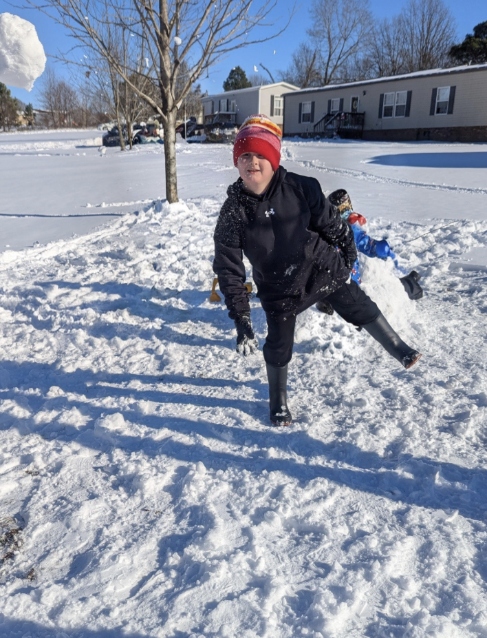 A child in a red and yellow knit hat runs through a snowy neighborhood yard while another child plays near a snowman in the background.