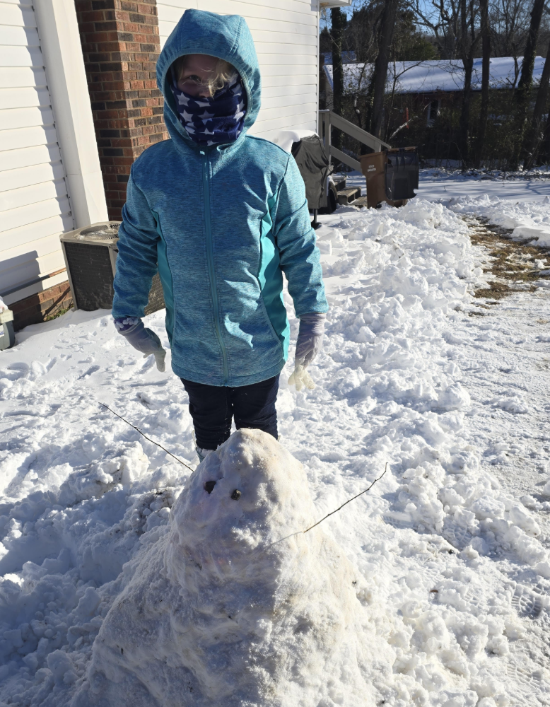 A child wearing a blue winter jacket and star-pattern face covering stands behind a small, rounded snowman with stick arms in a snowy driveway beside a house.
