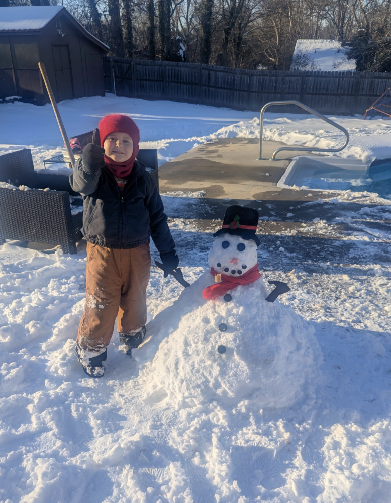 A child stands beside a snowman near a backyard swimming pool. The snowman wears a black top hat, red scarf, and has button details, while the child holds a stick and wears winter coat, pants, and a red hat.