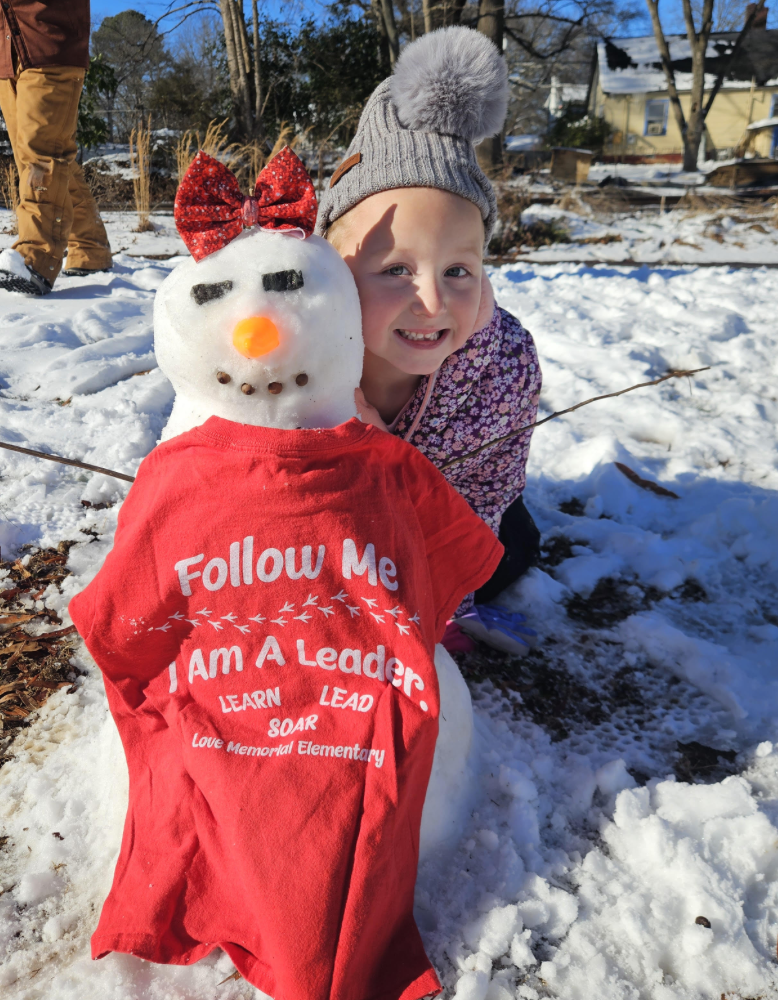 A child kneels beside a small snowman dressed in a red T-shirt that reads “Follow Me, I Am A Leader, Love Memorial Elementary.” The snowman has a red bow, carrot nose, and pebble smile.