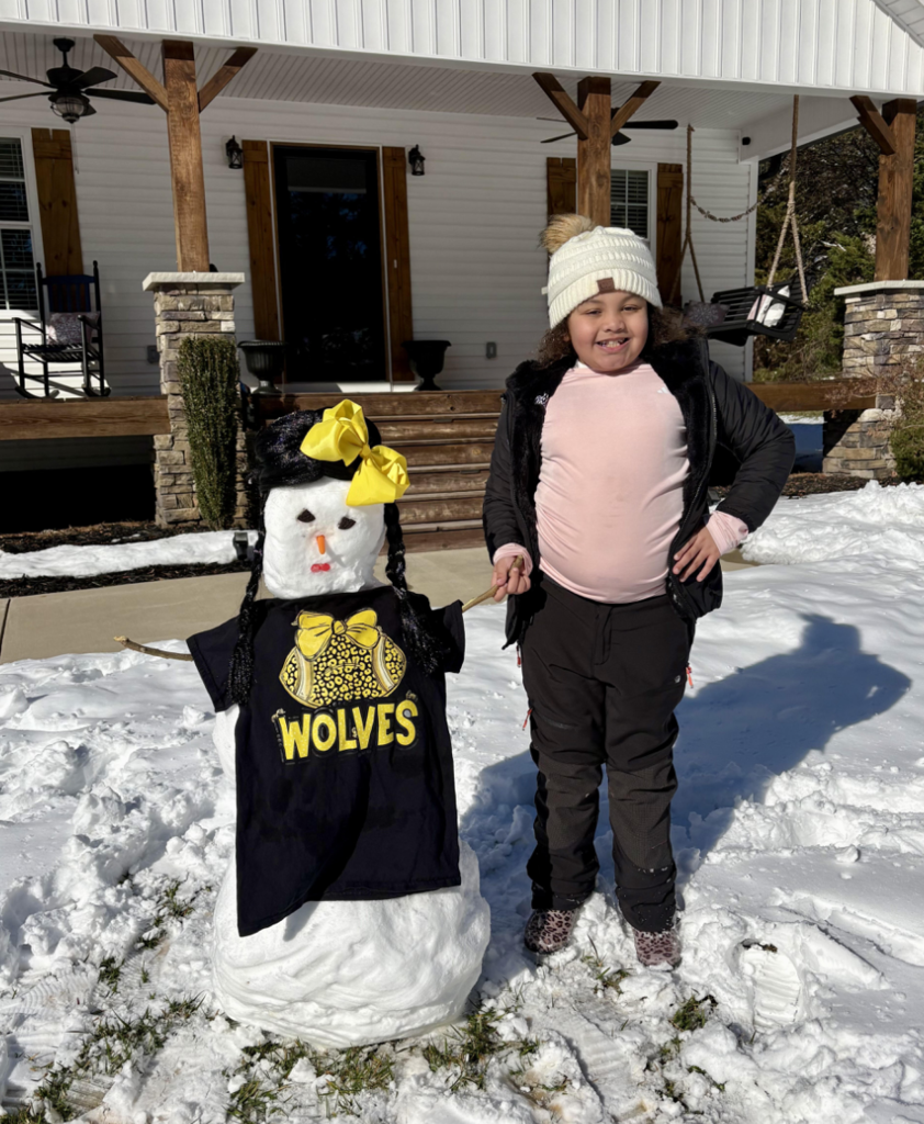 A child stands beside a snowman in front of a home porch on a sunny winter day. The snowman is dressed in a black Wolves T-shirt with a yellow bow and has braided yarn hair, a carrot nose, and stick arms. The child wears a white knit hat, pink shirt, and winter jacket, smiling with one hand on their hip.