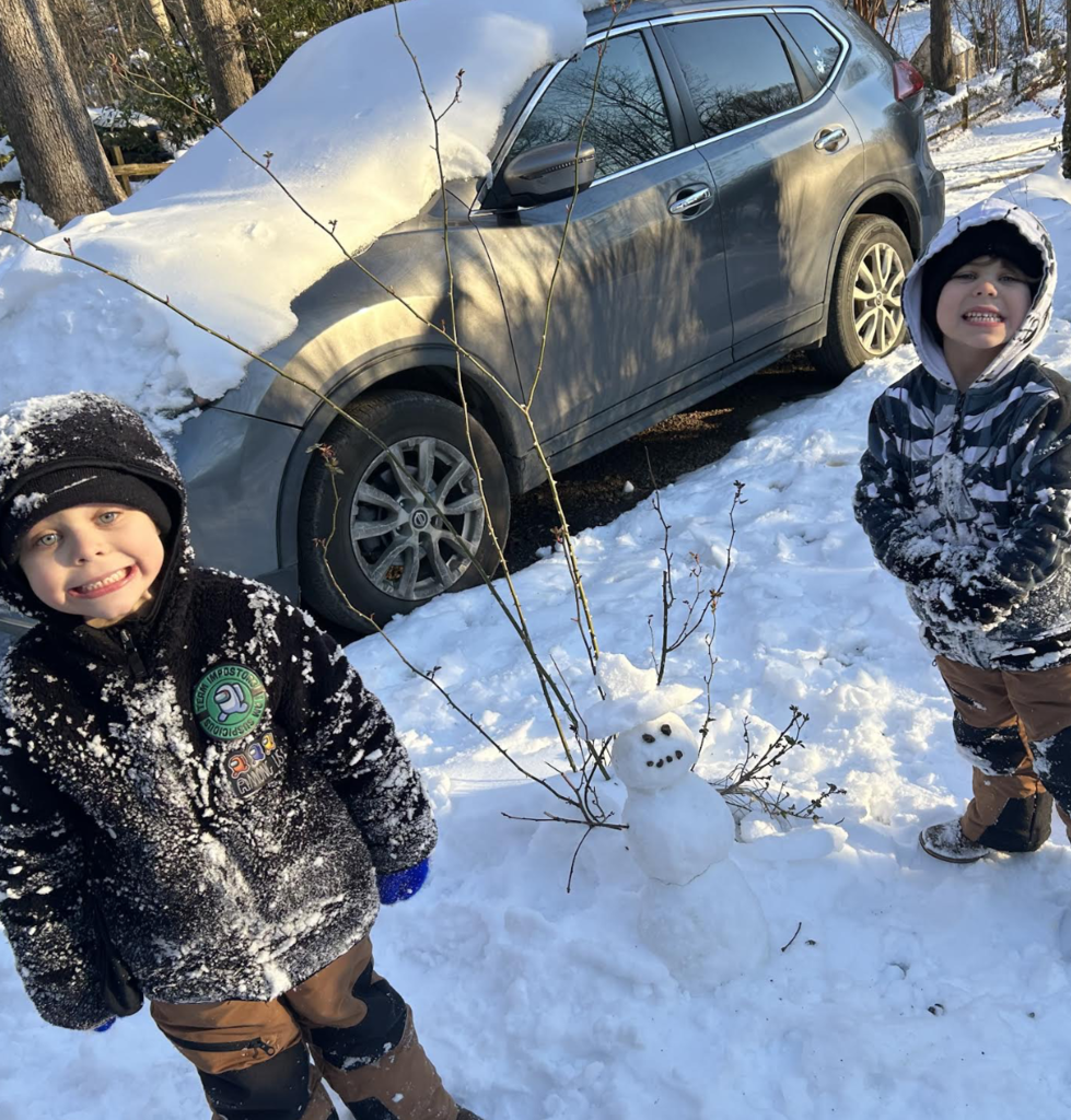 A child stands next to a snowman dressed in a Wolves shirt with a yellow bow and braided yarn hair. They pose in front of a home porch on a sunny winter day.