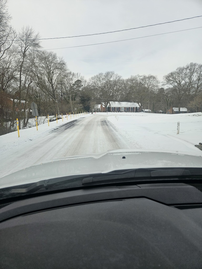 View from inside a vehicle traveling along a rural road covered with snow and ice. The roadway shows faint tire tracks with yellow reflective posts lining the left shoulder. A snow-covered yard and a brick home with a white roof appear ahead, with bare winter trees and an overcast gray sky surrounding the scene.