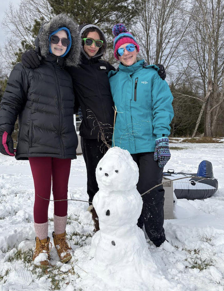 Three students in colorful winter coats and hats pose together behind a small snowman they built, standing in a snowy yard with sleds nearby.
