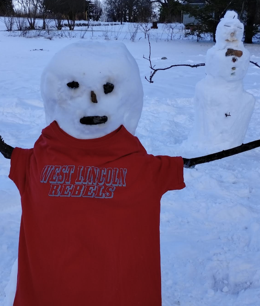 A snowman wearing a red “West Lincoln Rebels” T-shirt stands in a snowy field with stick arms stretched wide and another snowman in the background.